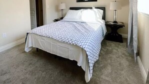 A bedroom with a bed, two bedside tables, a nightstand, a ceiling light, a door, and a window with curtains. - Alaska Townhouse Getaway: Anchorage Cozy Retreat - Close to Downtown (Anchorage)