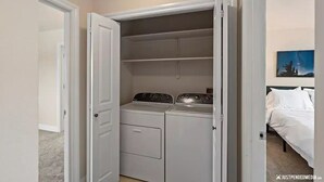 A laundry room with two washing machines and a dryer, a bed with white bedding, and a painting on the wall. - Alaska Townhouse Getaway: Anchorage Cozy Retreat - Close to Downtown (Anchorage)
