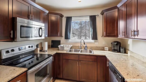 A kitchen with dark wooden cabinets, stainless steel appliances, a granite countertop, a window with curtains, and a coffee maker. - Alaska Townhouse Getaway: Anchorage Cozy Retreat - Close to Downtown (Anchorage)