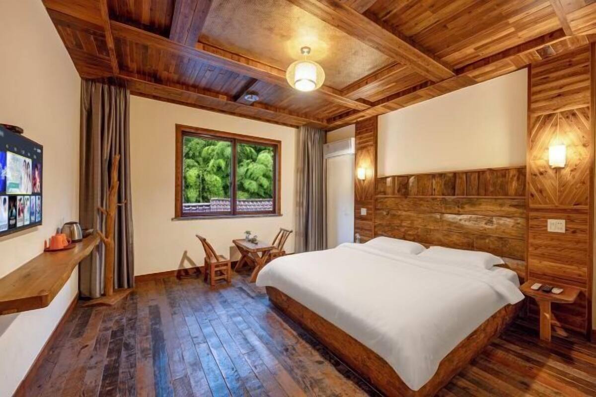 A wooden-paneled room with a bed, a TV, a window with a view of greenery, and a small table with chairs.