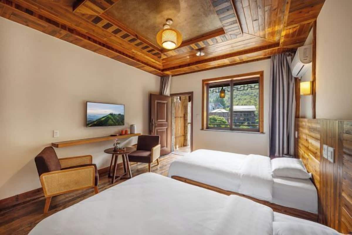A hotel room with wooden ceilings, two beds, a TV, a window with a view, and a small table with chairs.