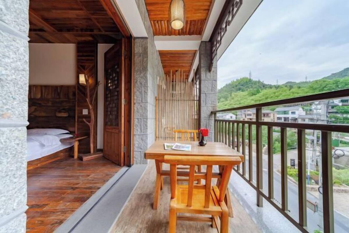 A balcony with a wooden table and chairs, a bed in the adjacent room, and a view of a green hillside and buildings.