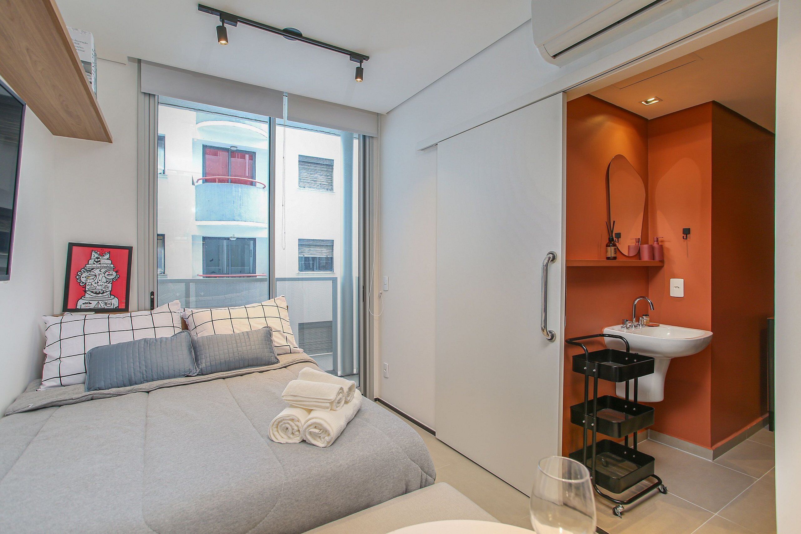 Ein Schlafzimmer mit Bett, Nachttisch, Fenster und einem Badezimmer mit Waschbecken und Spiegel.