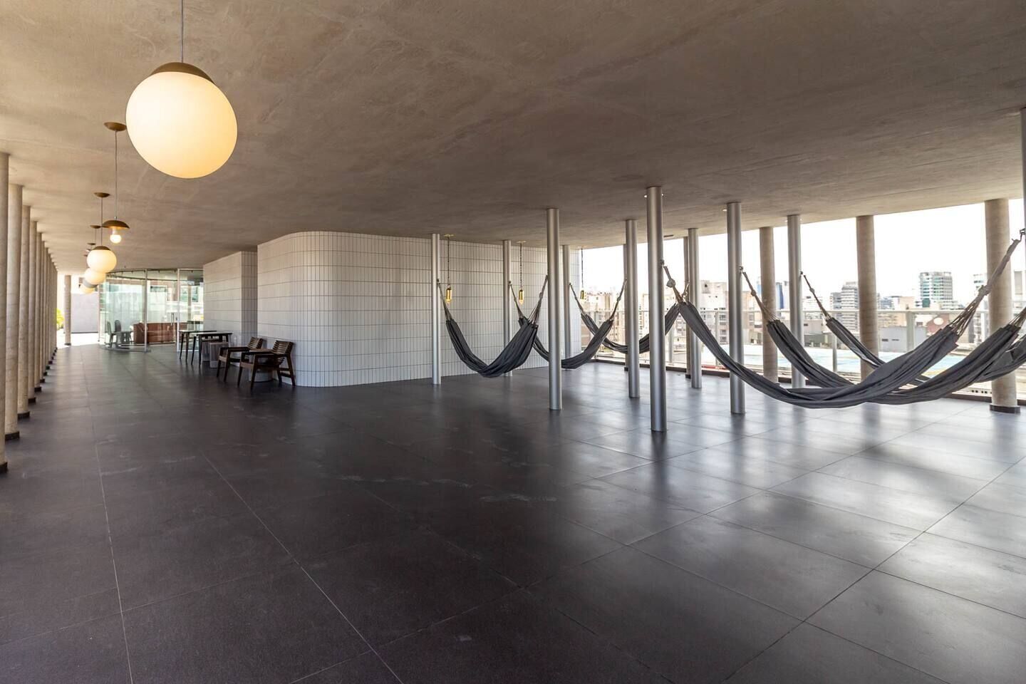 A spacious indoor area with a modern design, featuring a long row of hammocks and a seating area with tables and chairs.