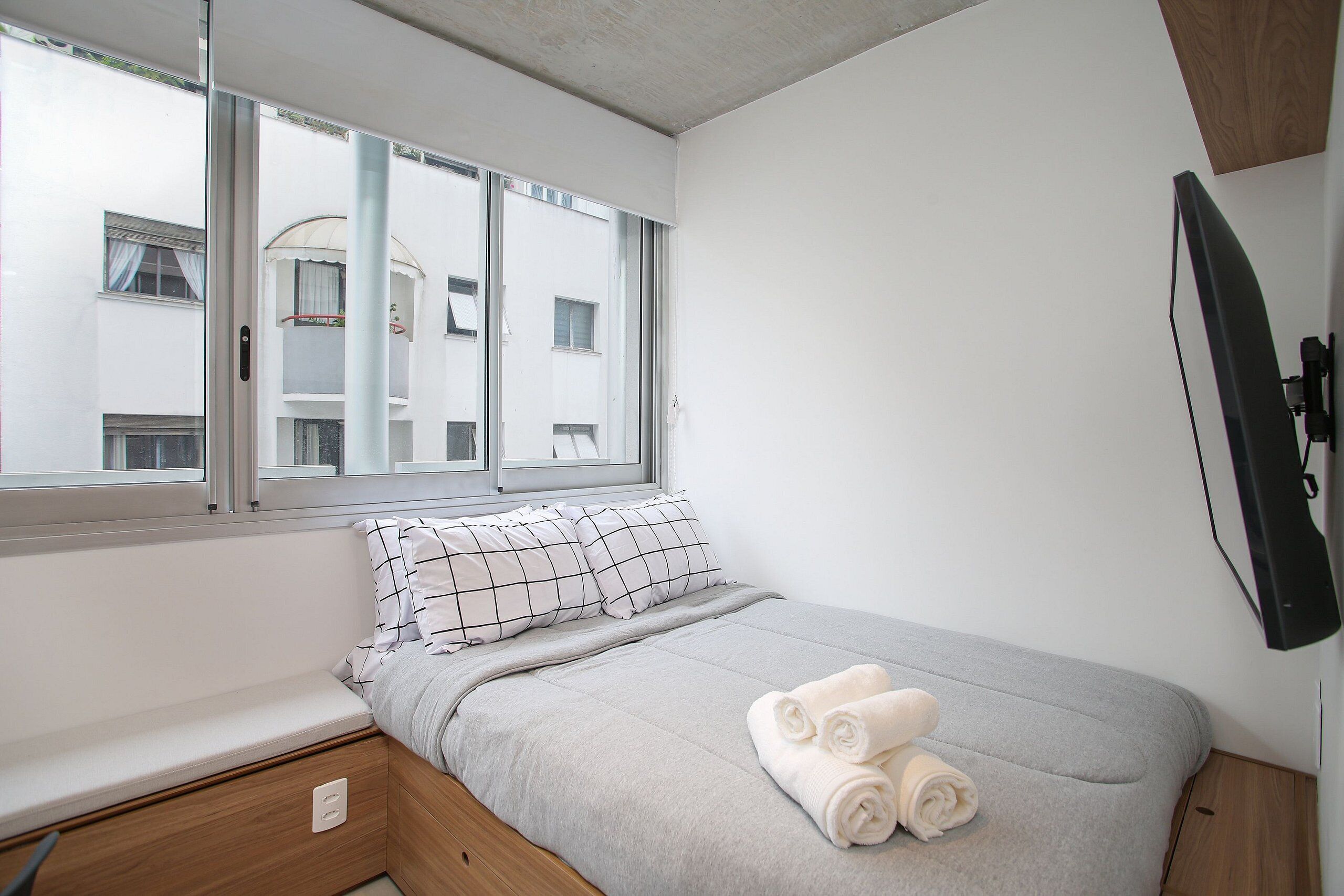 A single bed with a grey bedspread, two pillows, and rolled towels on the side. A window with a view of buildings is visible.