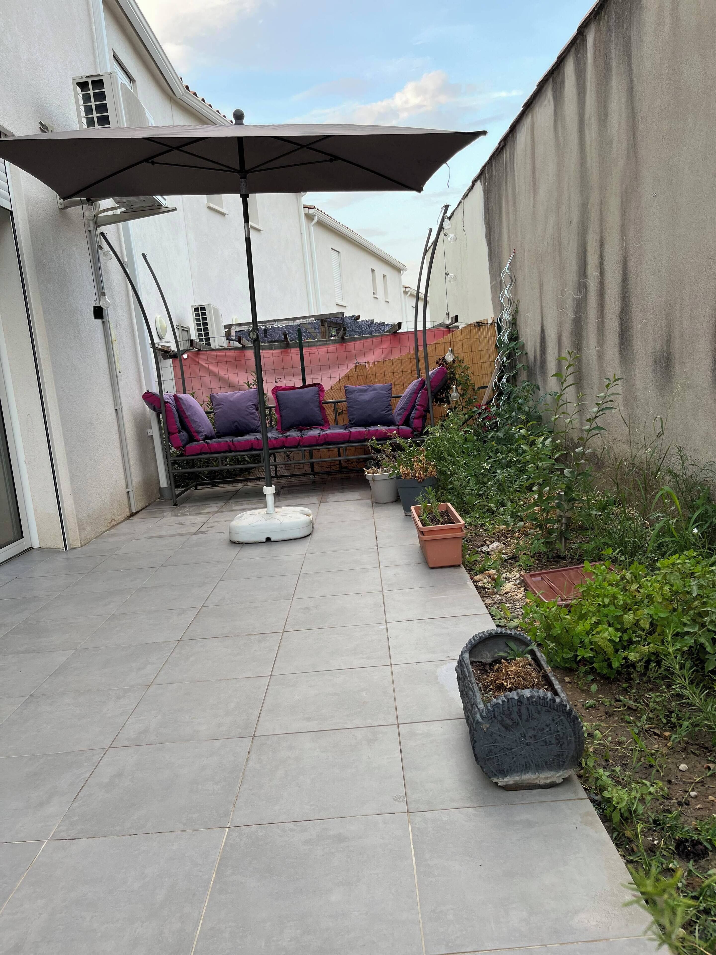 Un espace patio aménagé avec des sièges confortables, un parasol, des plantes en pot et un sol carrelé.