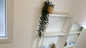 Et badeværelse med en hvid vask, et spejl, et hvidt reolsystem med kurve, en mønstret måtte og en potteplante.