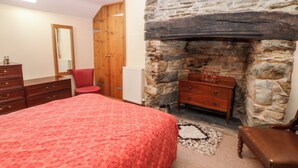 A bedroom with a stone fireplace, a wooden wardrobe, a red chair, and a bed with a red bedspread. - Ereiniog (Garndolbenmaen)