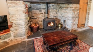 A stone fireplace with a wood-burning stove, a wooden table, and a patterned rug in a room with stone walls. - Ereiniog (Garndolbenmaen)