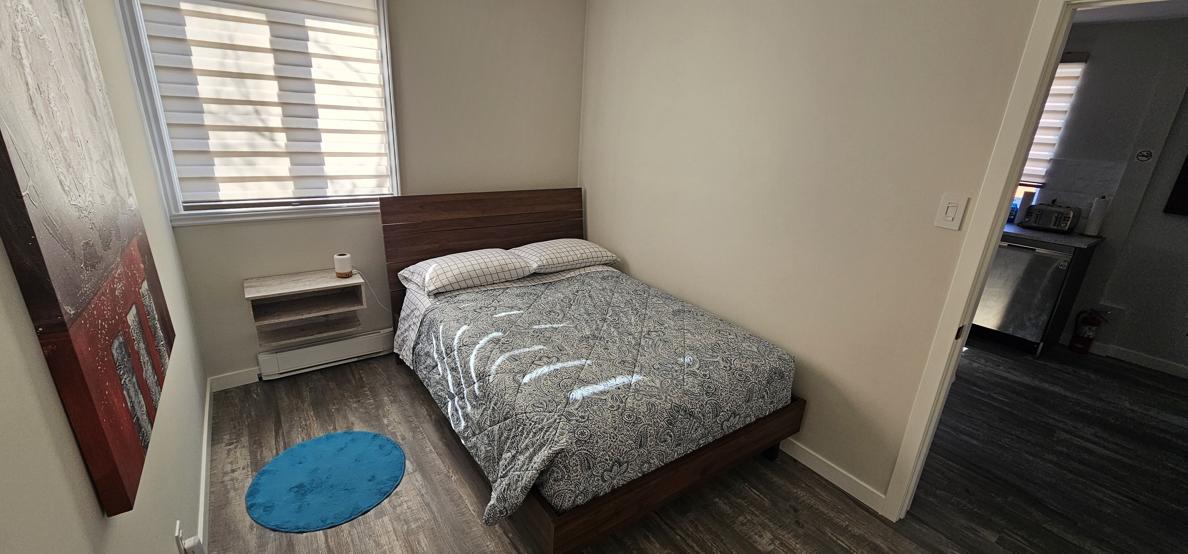 Un dormitorio pequeño con cama, ventana con persianas, alfombra azul y puerta que da a una zona de cocina.
