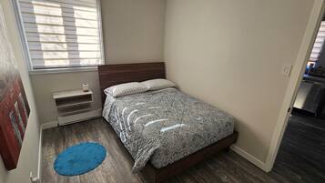 Un dormitorio pequeño con cama, ventana con persianas, alfombra azul y puerta que da a una zona de cocina.