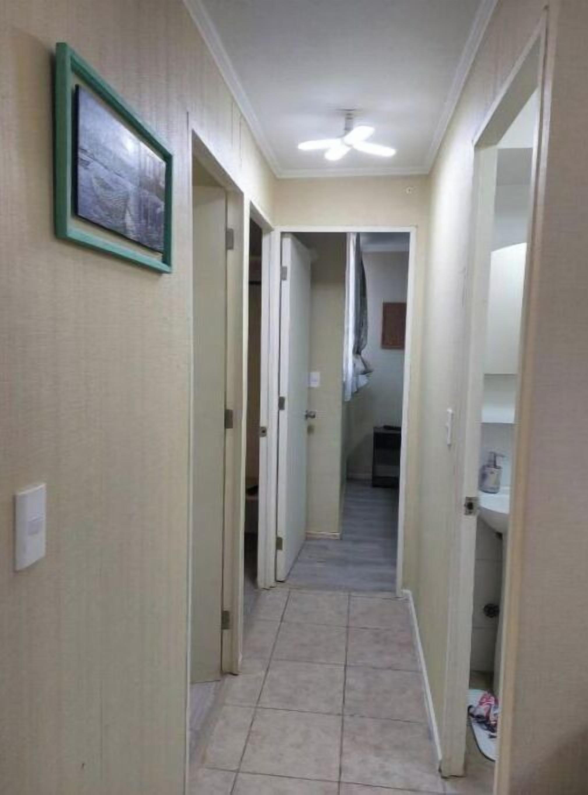 Un pasillo estrecho con un ventilador de techo, un cuadro en la pared izquierda y un baño al final.