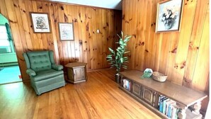 A room with wooden panel walls, a green armchair, a small cabinet, a potted plant, and a wooden bench with books. - Creekside Rural Retreat in Beautiful Ashe County
 (Lansing)