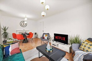 Ein modernes Wohnzimmer mit Essbereich, einem Flachbildfernseher, der 'NETFLIX' anzeigt, und einer Uhr an der Wand.