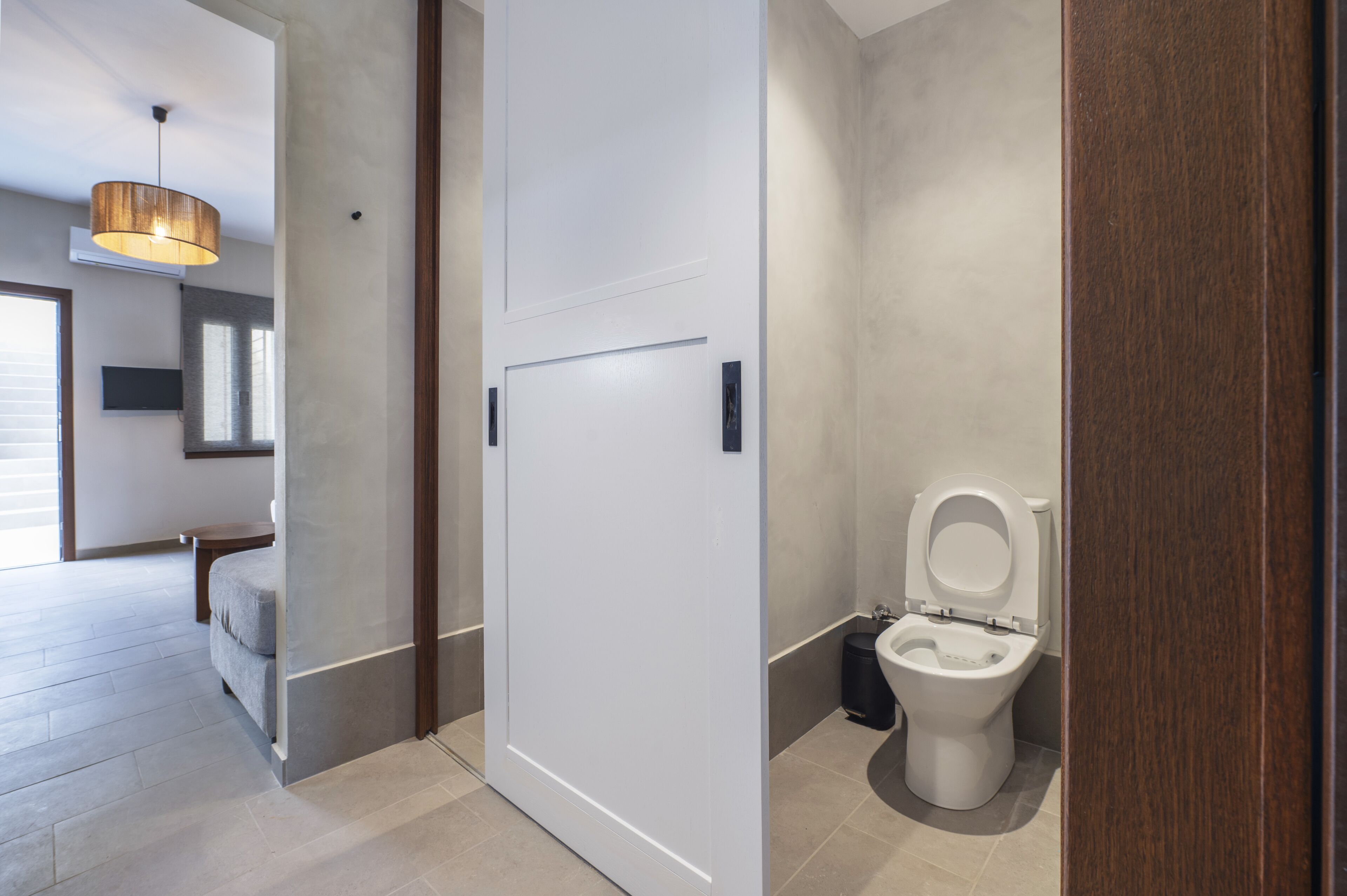 A modern bathroom with a toilet, a sink, and a trash bin. There is a wooden door and a light fixture on the ceiling.