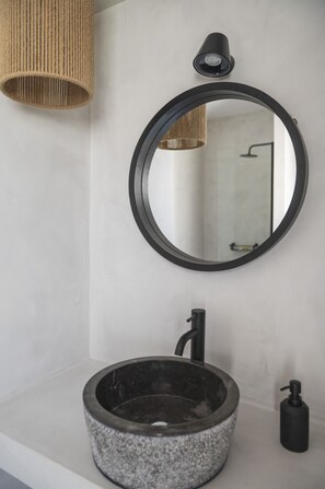 A bathroom with a round mirror, a stone sink, a black faucet, a black soap dispenser, and a woven light fixture. - Casa di Demezzo (Sitia)