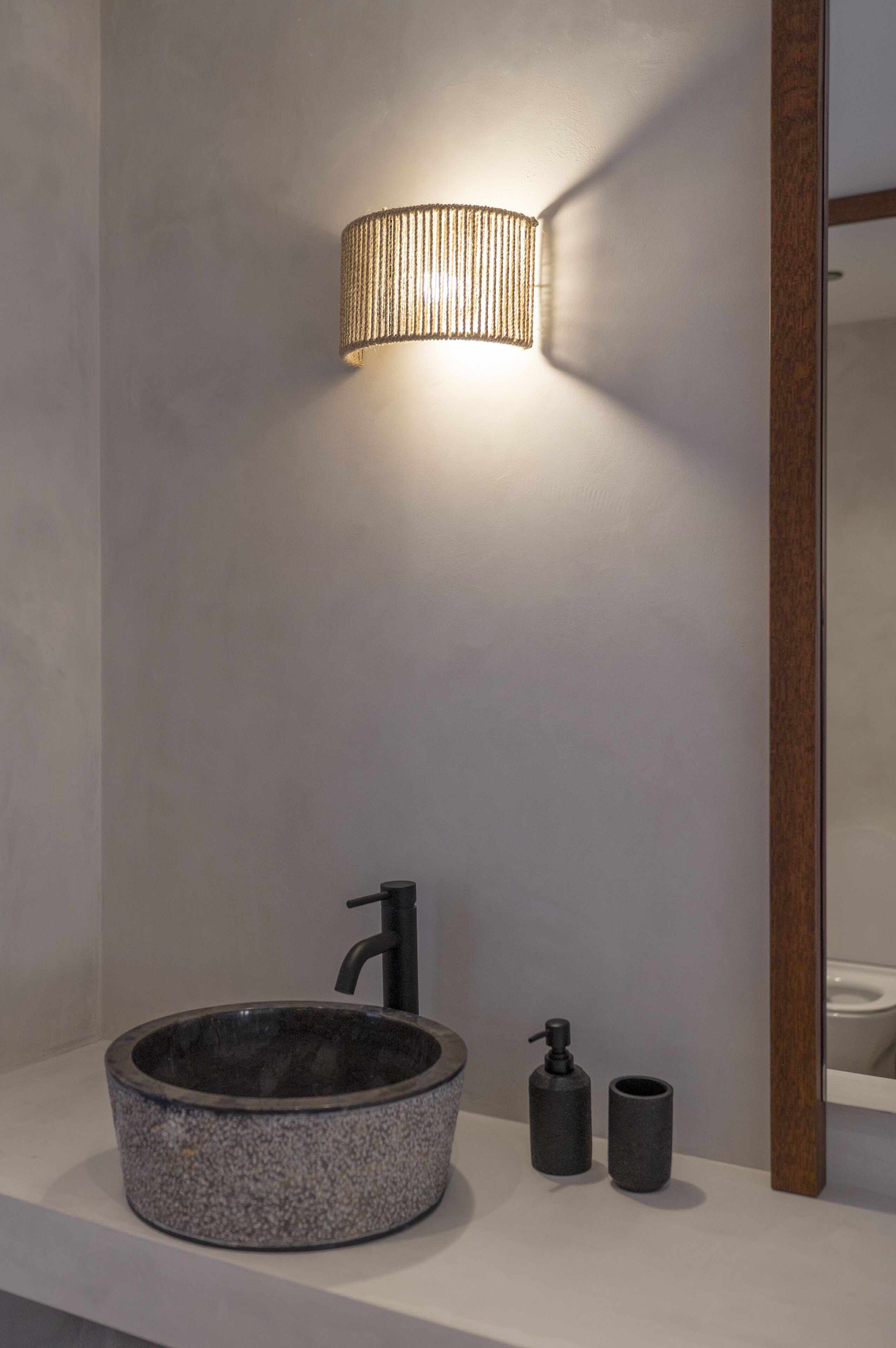 A modern bathroom with a round stone sink, a black faucet, a wall-mounted light fixture, and a small mirror.