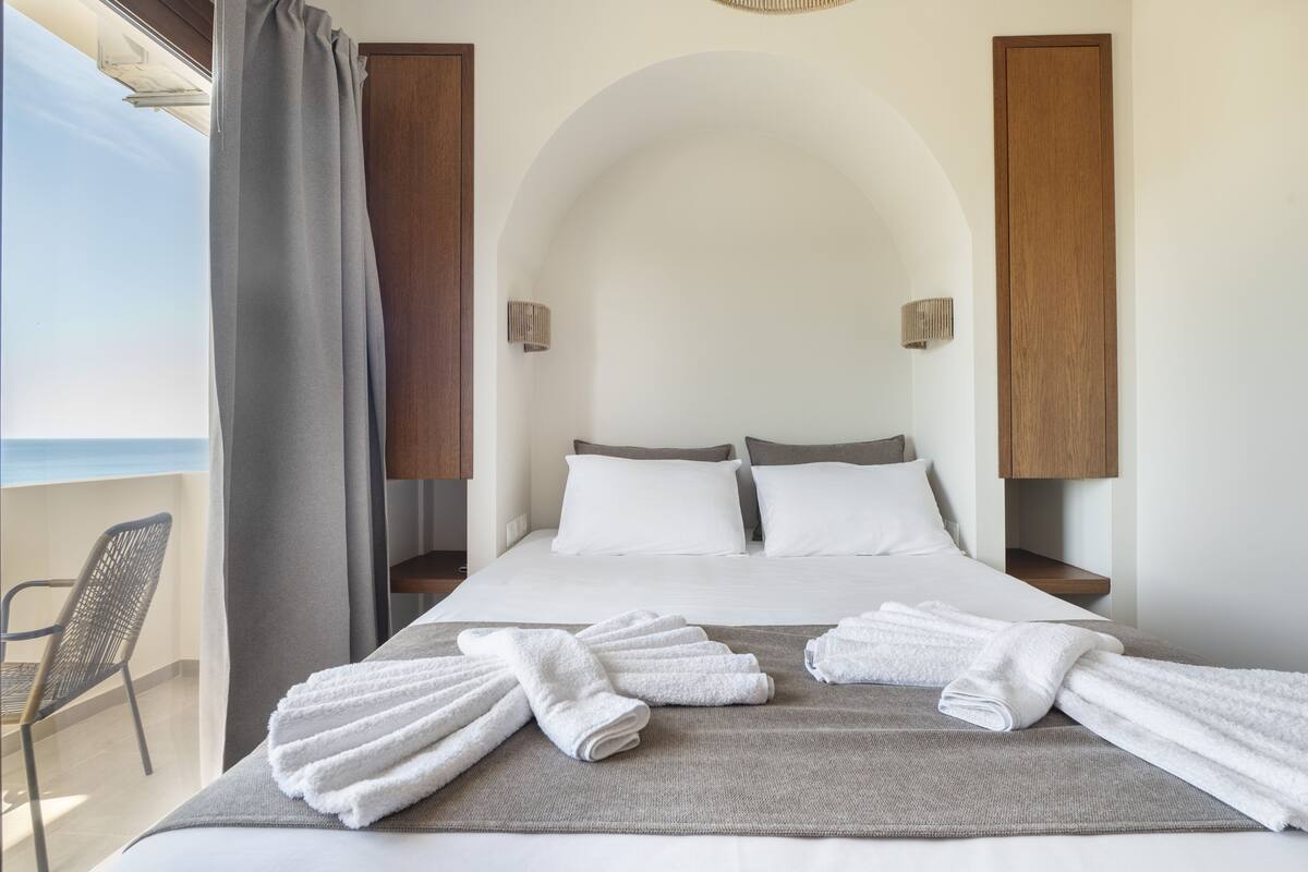 A hotel room with a bed, towels, a chair, and a view of the sea.