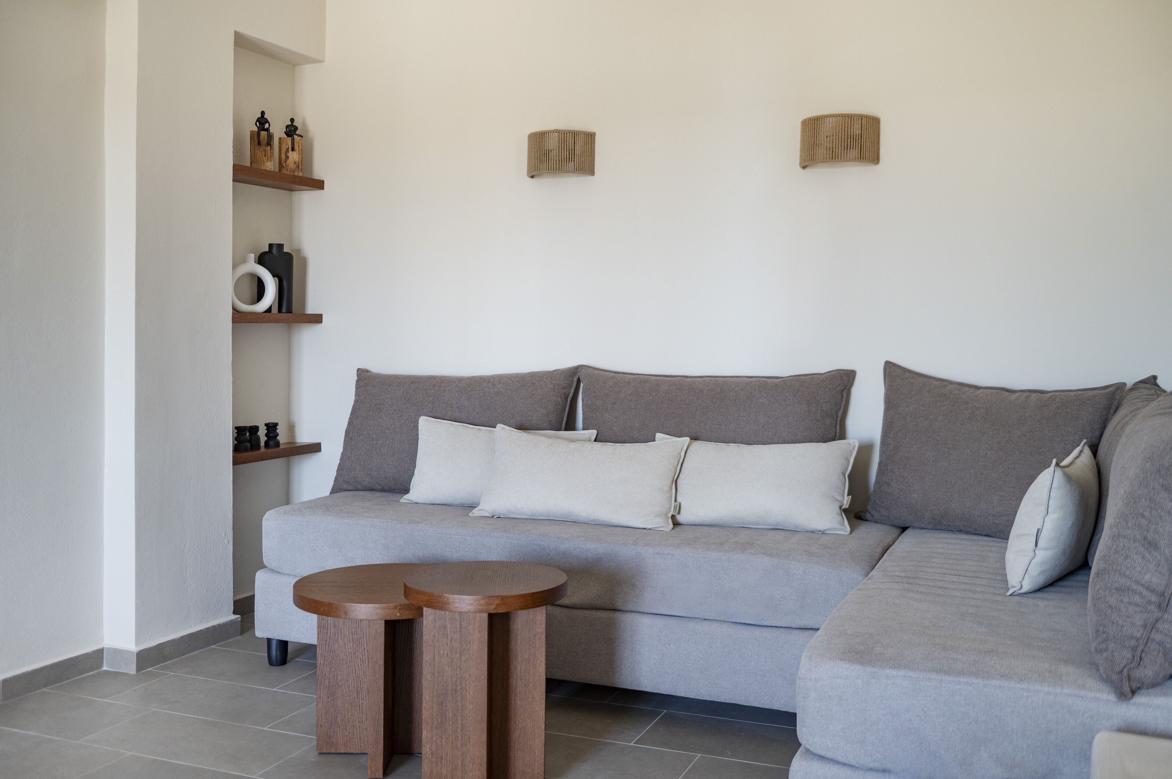 A modern living room with a grey sofa, two wooden side tables, and a white wall with shelves holding decorative items.
