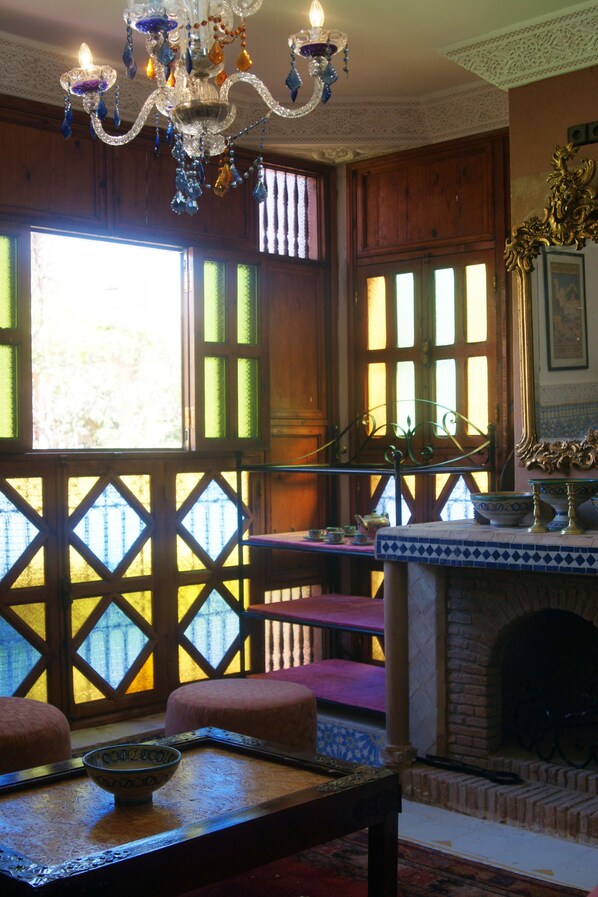 A room with a chandelier, wooden paneled walls, a fireplace, and a view through a lattice window.