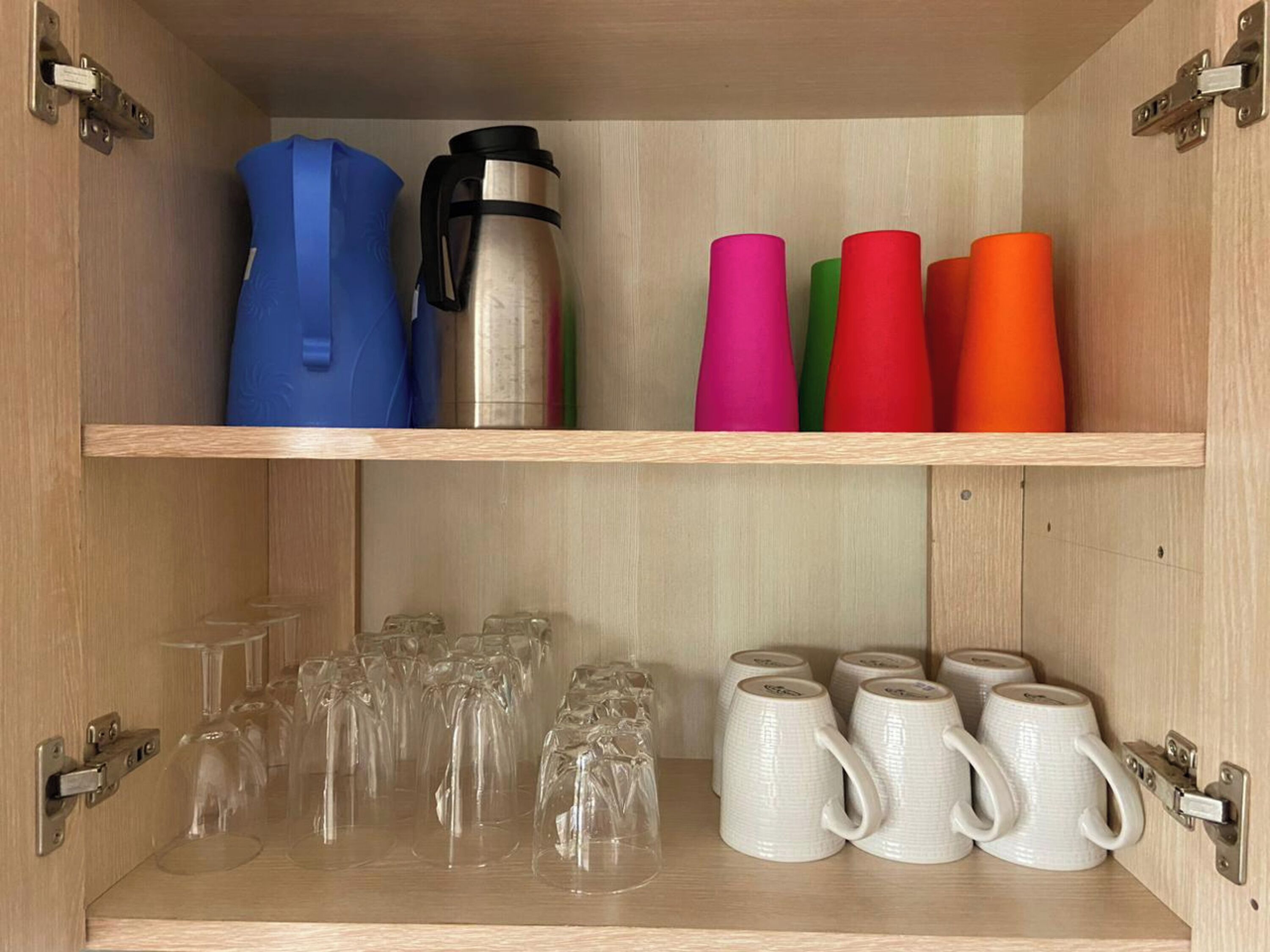 A wooden cabinet with shelves holding a stainless steel water bottle, colorful cups, and clear glassware.