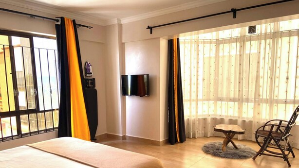 A hotel room with a bed, a TV, a small table, a chair, and a view of the outside through large windows. - Midtown B61 Apartment (Nakuru)