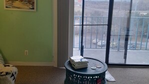 Uma sala de estar com uma janela ampla, um quadro na parede e uma mesa redonda com livros.