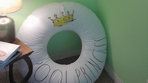Um quarto com parede verde, uma piscina insuflável branca com coroa e a inscrição 'POOLPRINCE', uma lâmpada e um livro sobre uma mesinha.