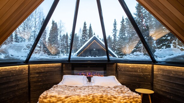 Ein gemütliches Blockhauszimmer mit einem großen Bett, einem Kamin und Blick auf einen verschneiten Wald.