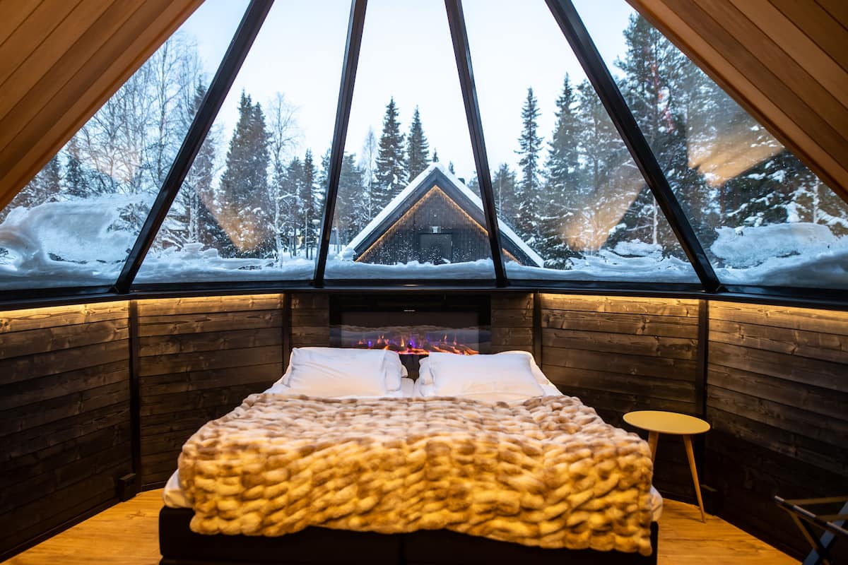 Habitació d'una cabana acollidora amb un llit gran, una llar de foc i vista a un bosc nevat.