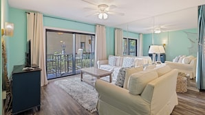 A living room with a sofa set, a coffee table, and a TV. There is a balcony with a view of greenery. - Surfside Hideaway (Hilton Head Island)