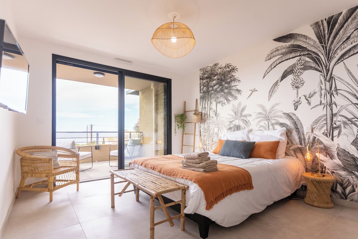 Quarto com cama, um banquinho, cadeira, mesa de cabeceira, luminária, painel na parede e varanda com vista.