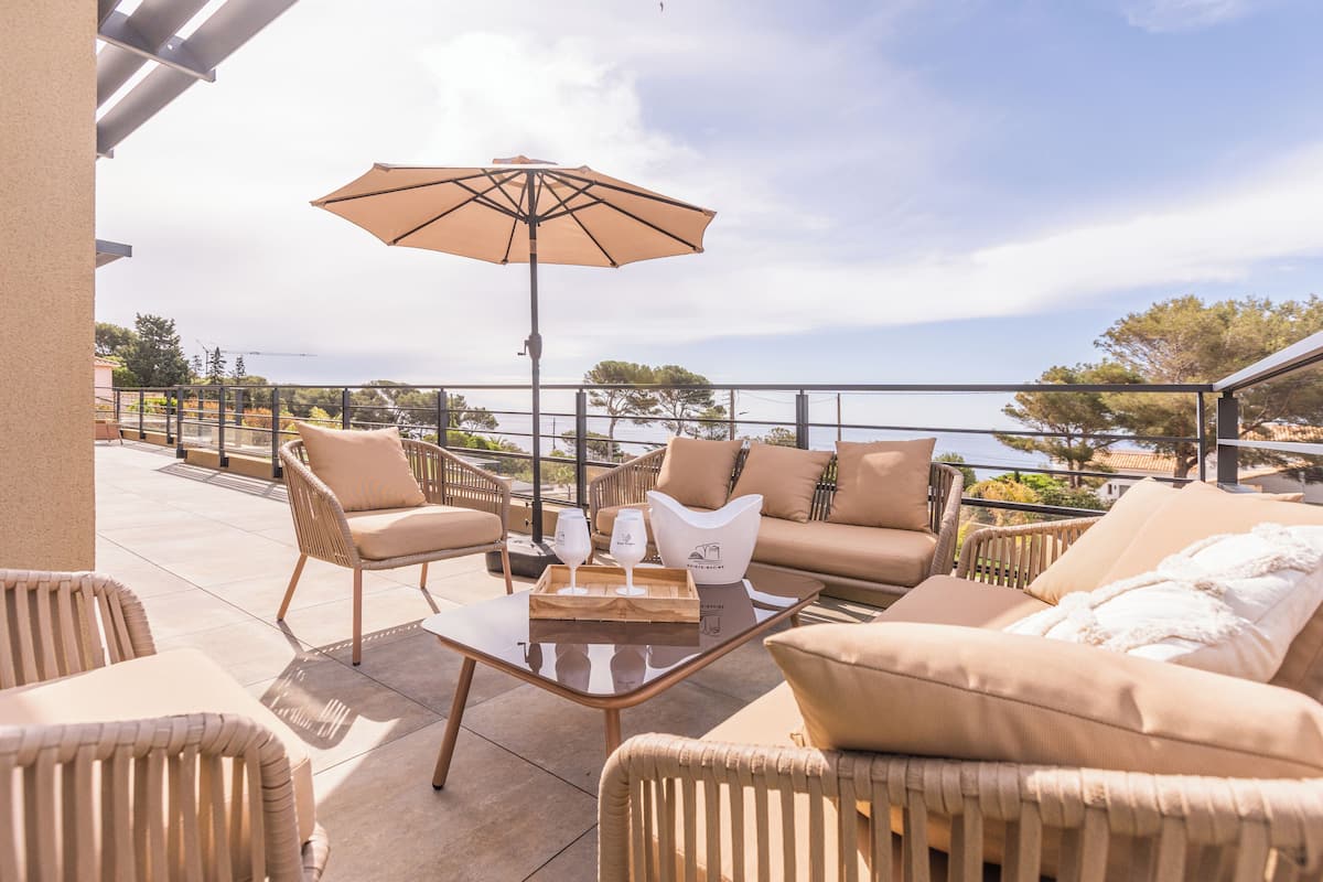 Uma área externa ensolarada com mobiliário de vime, uma mesa de vidro e um grande guarda-sol.
