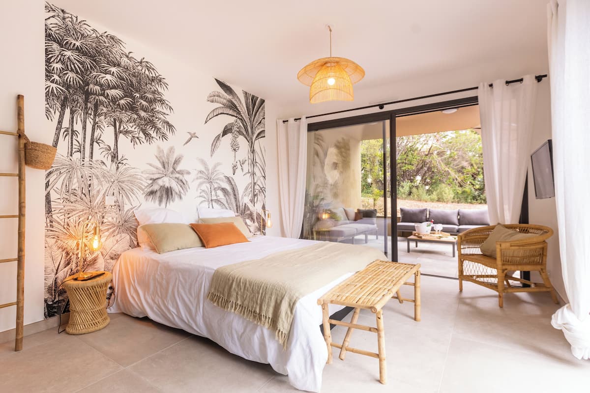 Quarto com uma cama grande, mesa de cabeceira com luminária, painel decorativo na parede e vista para o exterior por uma porta de correr.
