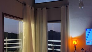 Une pièce dotée d’une grande porte-fenêtre coulissante donnant sur un balcon, d’un téléviseur fixé au mur et d’une table avec une lampe.