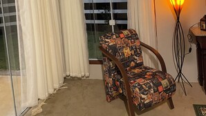 Une pièce avec une chaise près d’une fenêtre ornée de rideaux blancs, un lampadaire et une vue sur un balcon.