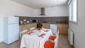A modern kitchen with white appliances, a dining table set for four with orange chairs, and a view of the outside through a window. - Logis de la Perruche - Maison avec terrasses (Montreuil-Bellay)