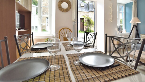 A dining table set with plates, glasses, and a mat. There are chairs around the table, and a window with a view of a building outside. - Mézange Bleue - Près du canal d'Ille et Rance (Lanvallay)