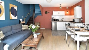 A compact living space with a sofa, coffee table, and dining table. A kitchen with modern appliances and a staircase is visible. - Mézange Bleue - Près du canal d'Ille et Rance (Lanvallay)