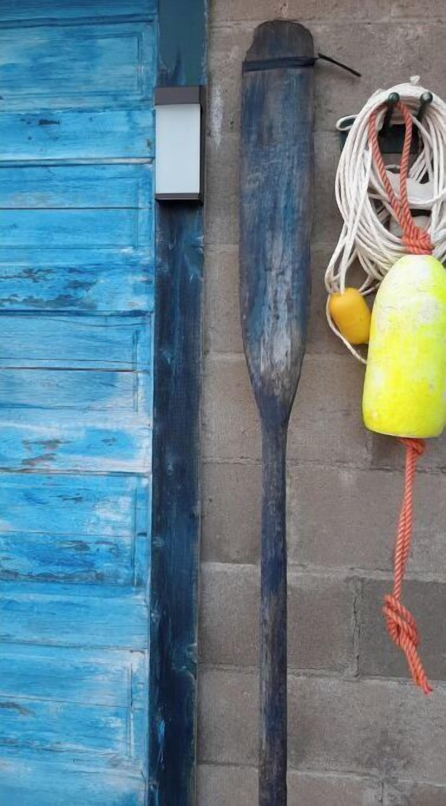 Two wooden paddles leaning against a brick wall, a blue door, and a yellow buoy with a rope.