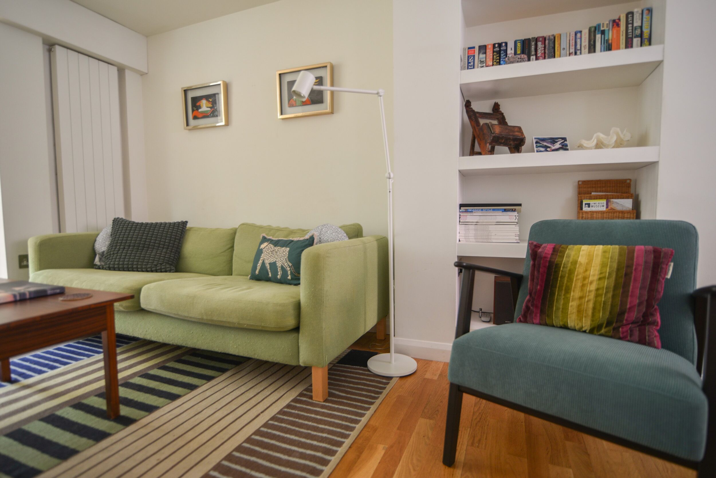 A living room with a green sofa, a teal armchair, a floor lamp, a bookshelf, and a striped rug.