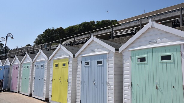 Une rangée de cabanes de plage colorées, chacune dotée d’une porte numérotée, un escalier en béton et une rambarde métallique en arrière-plan.