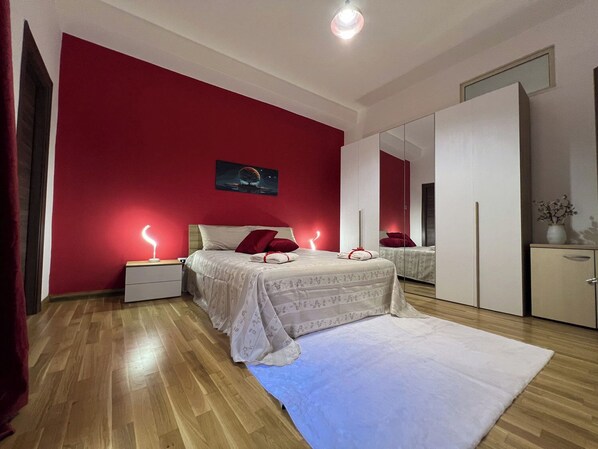 A bedroom with a red wall, a bed with white bedding, a nightstand, a bedside lamp, a mirror, and a wardrobe.