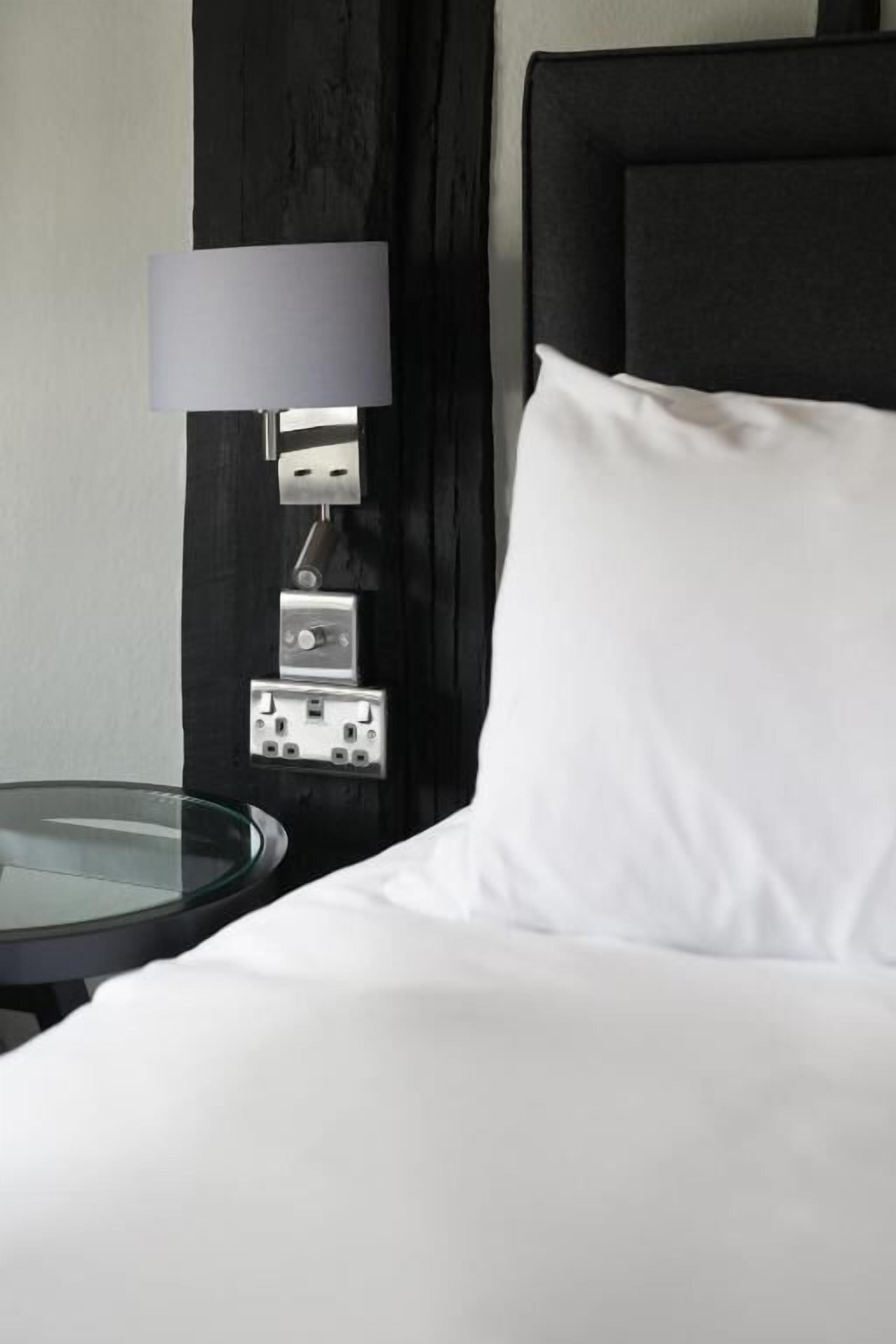 A bed with white bedding, a round glass table, a wall-mounted lamp, and a power outlet.