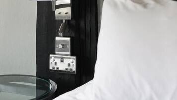 Un lit avec une literie blanche, une table ronde en verre, une lampe fixée au mur et une prise de courant.