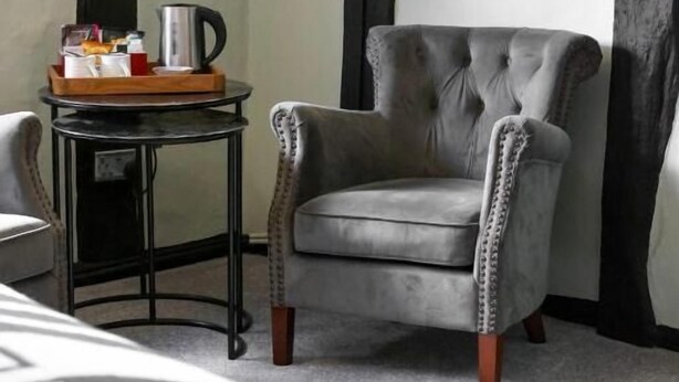 Una habitación con un sillón capitoné, una mesita con una bandeja de objetos y una cama con colcha estampada.