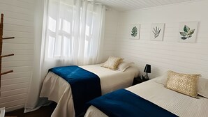 Ein Zimmer mit zwei Betten, weißen Wänden und einem Fenster mit transparenten Vorhängen.