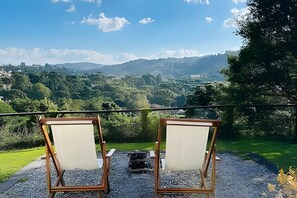 Two wooden chairs with white cushions on a gravel surface, overlooking a lush green landscape with hills and a clear blue sky. - Casa Blanca Campos do Jordão (Campos do Jordão)