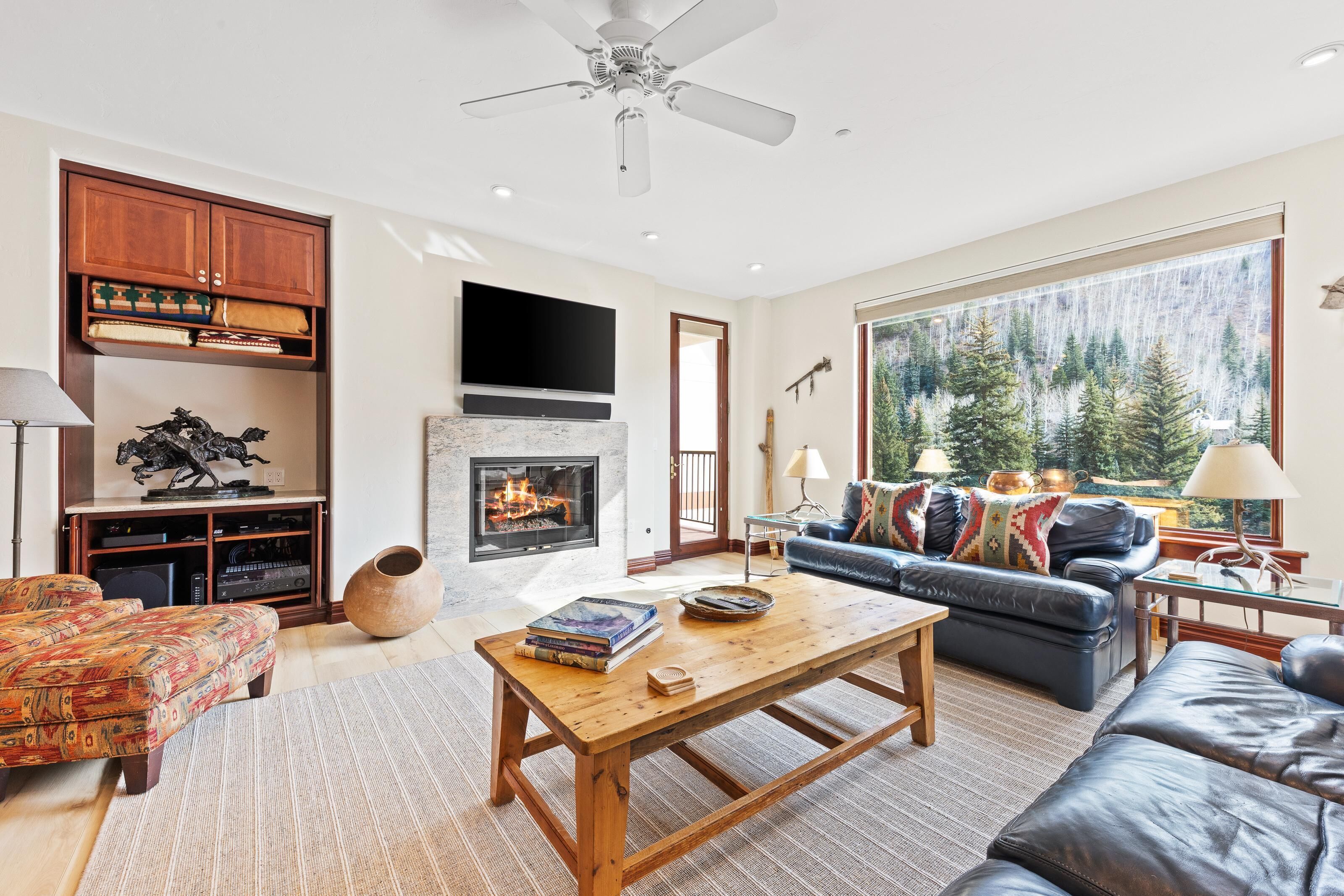 Una sala de estar acogedora con chimenea, un ventanal con vistas a un bosque, un sofá de cuero y una mesa de centro de madera.