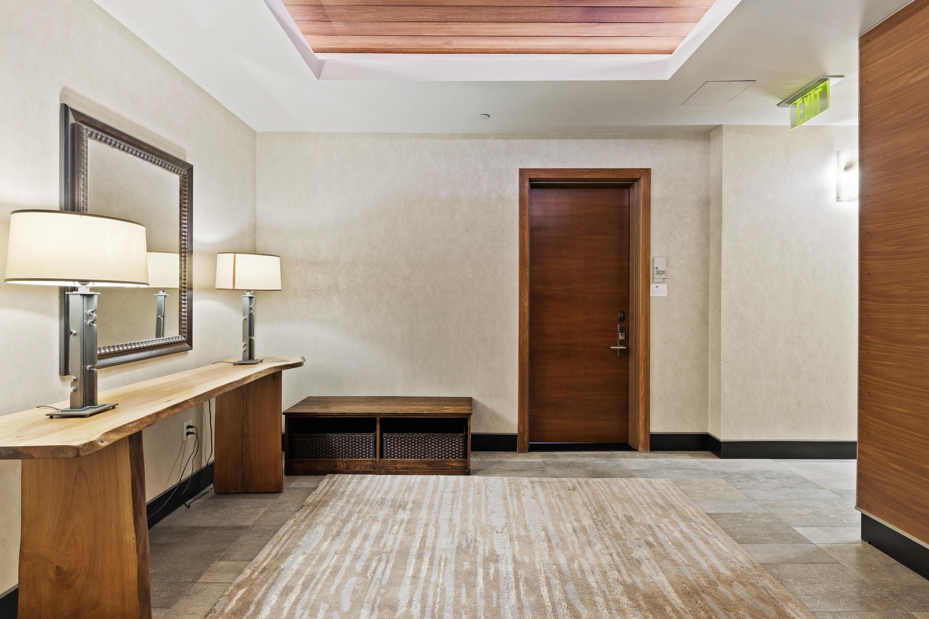 Un hall d’hôtel avec un comptoir d’accueil en bois, deux lampes fixées au mur, un banc, un grand miroir et une porte en bois.